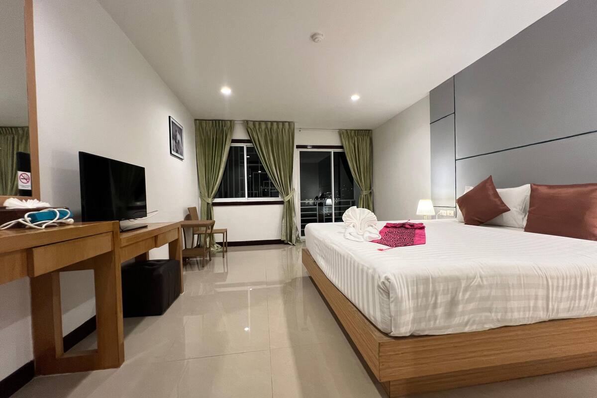 A hotel room with a bed, a desk with a TV, a chair, a window with curtains, and a night view outside.