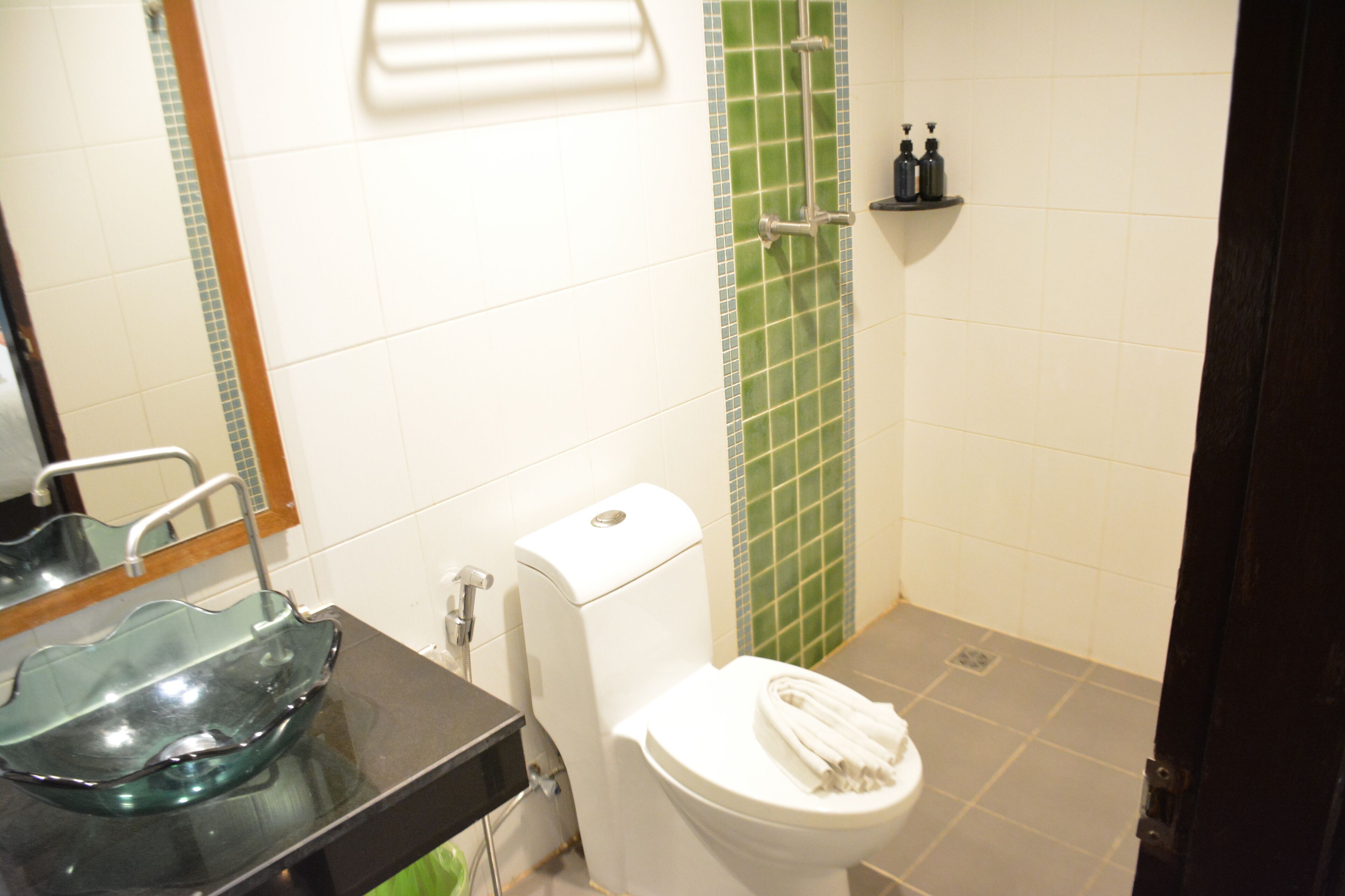 Un baño con un lavamanos de vidrio, un inodoro blanco y una zona de ducha con azulejos verdes.