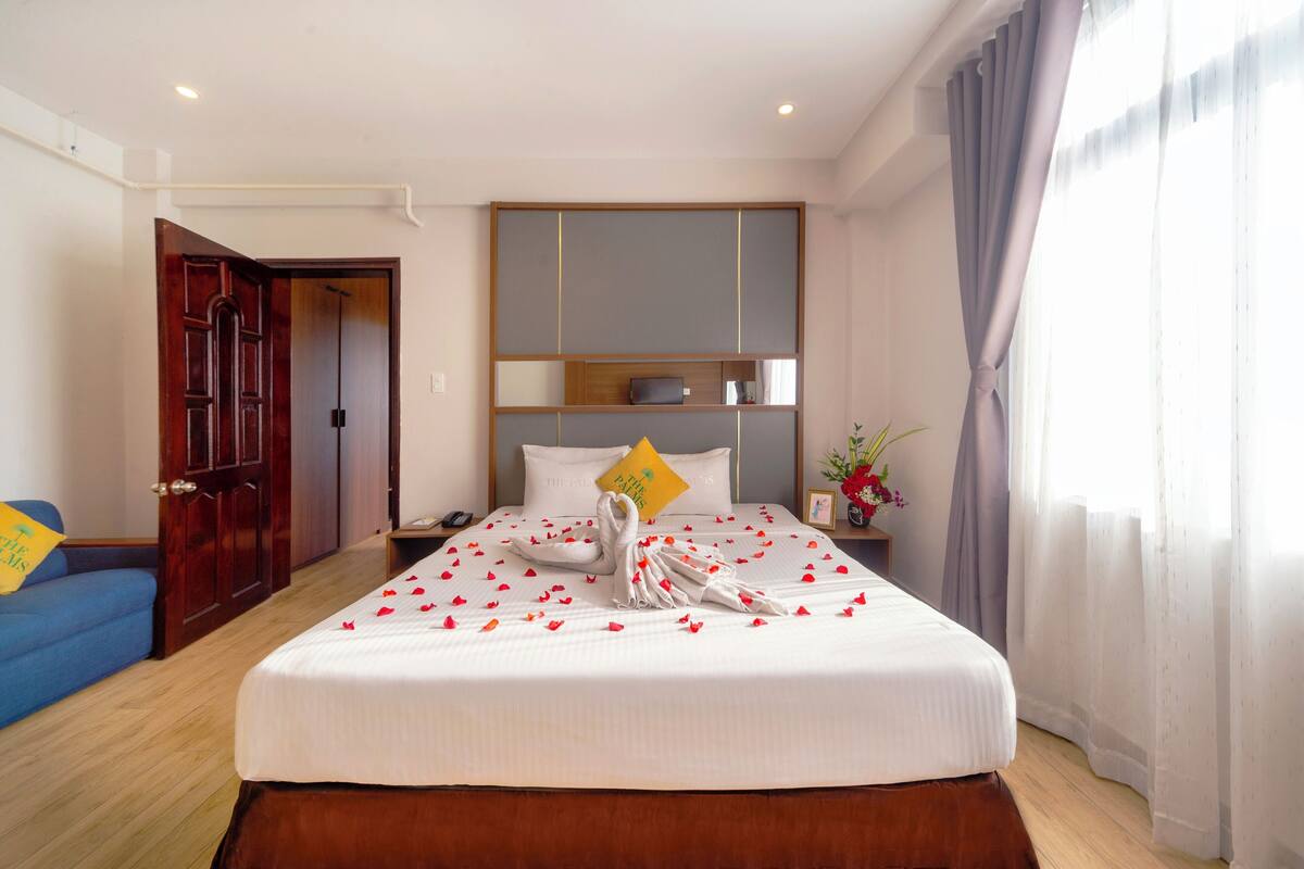 Une chambre d’hôtel avec un grand lit, une tête de lit en bois, une table de chevet, un bureau, un canapé bleu et une porte donnant sur une autre pièce.