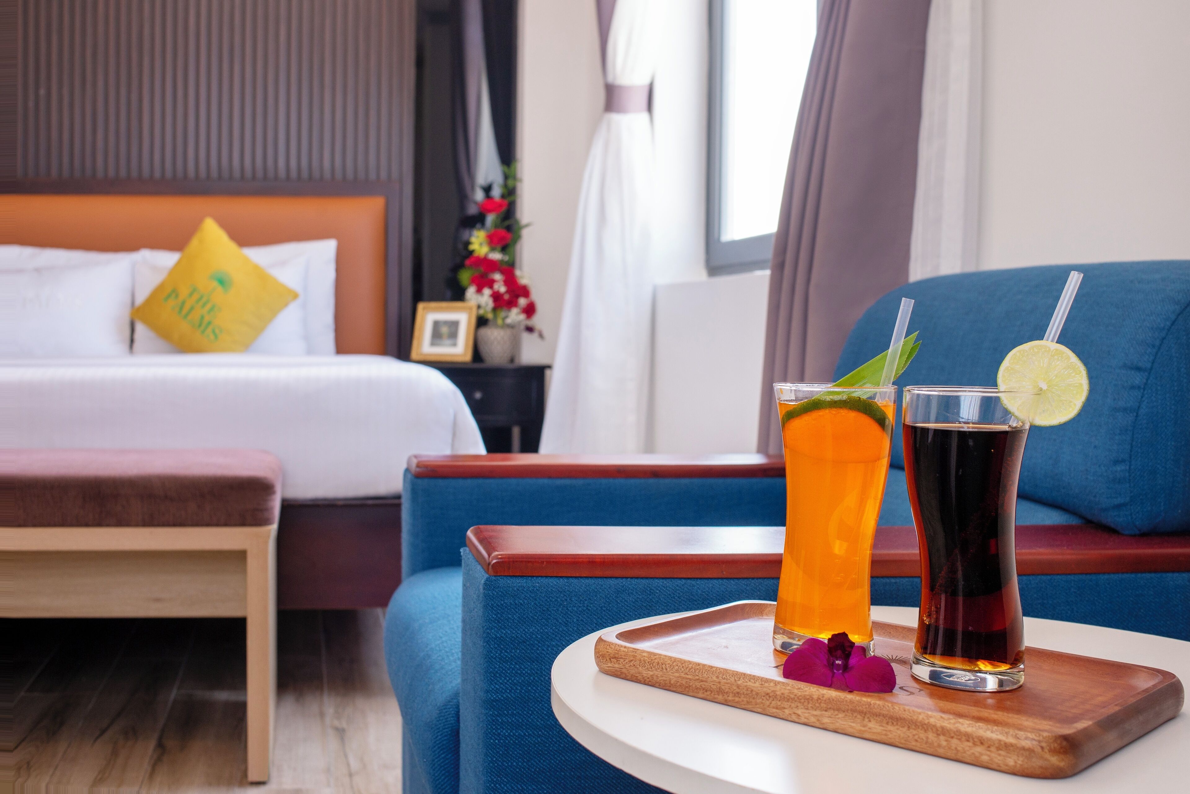 A hotel room with a bed, a blue armchair, a small table with drinks, and a nightstand with flowers.