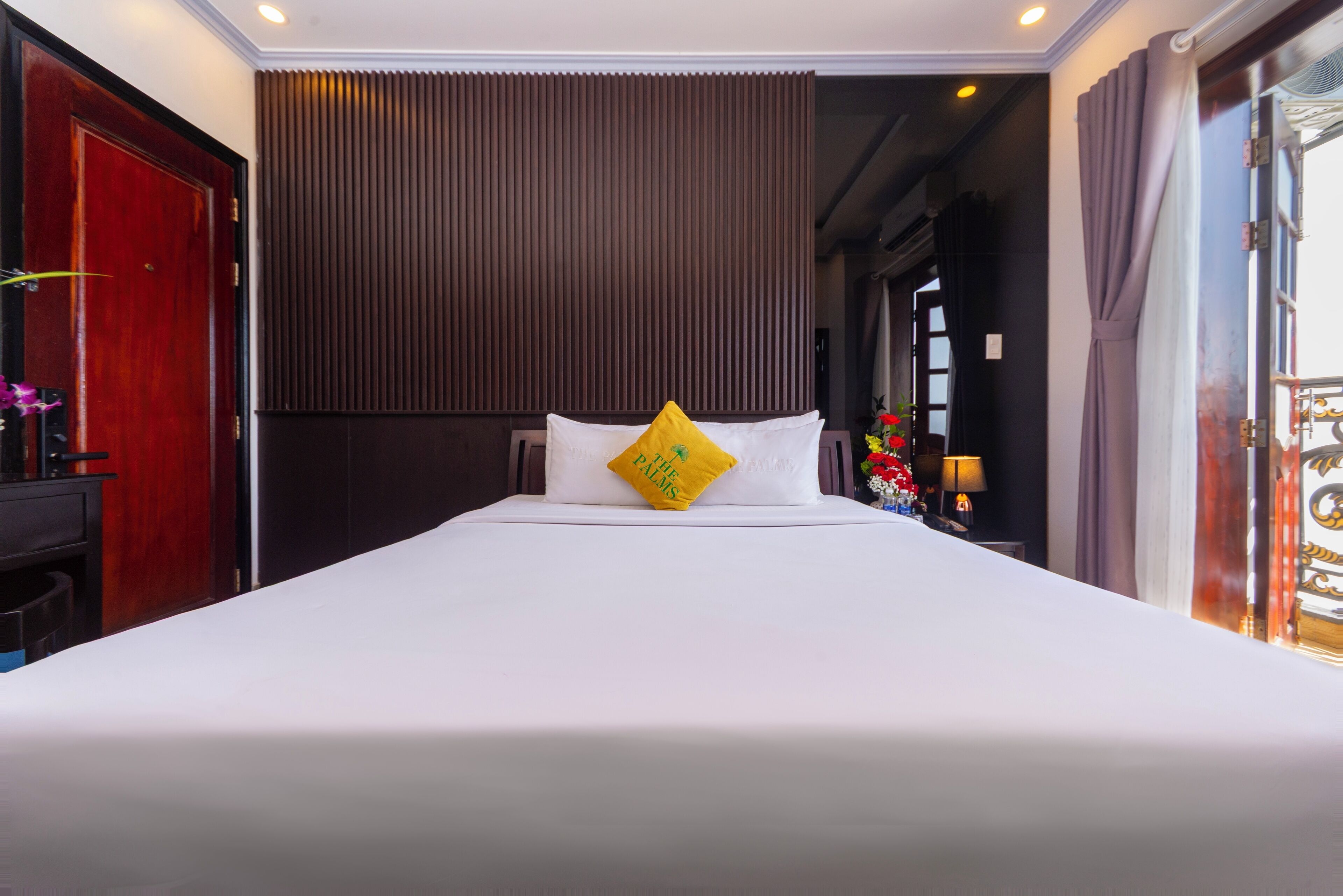 Una habitación de hotel con una cama grande, una puerta de madera, un panel de pared oscuro y una ventana con cortinas.