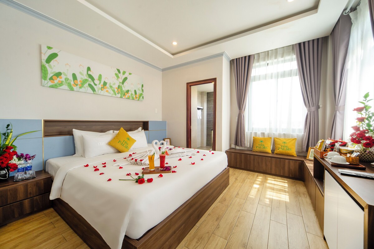 Una habitación de hotel con una cama grande, una mesita de noche con un jarrón de flores, una ventana con cortinas y un cuadro en la pared.