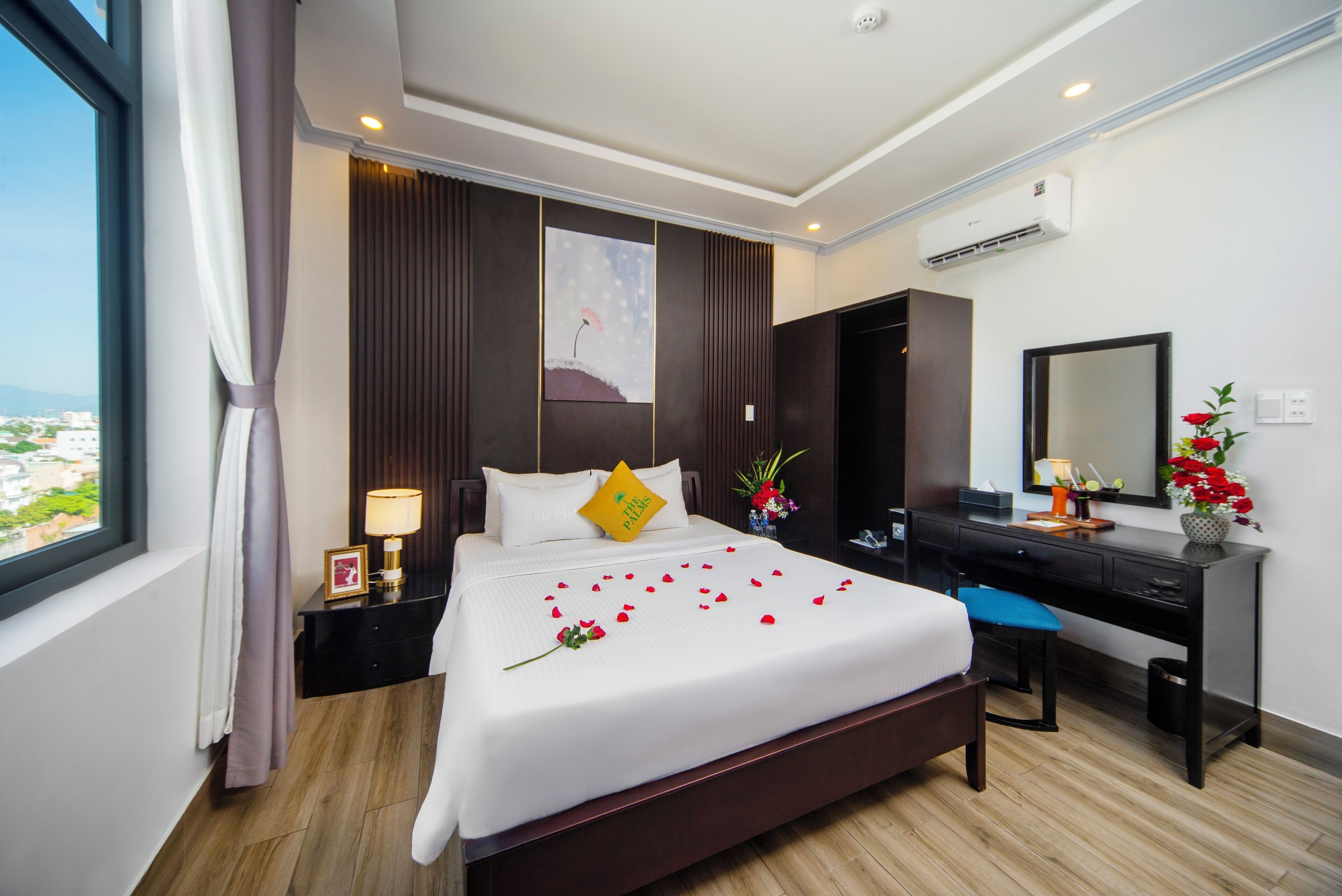 A hotel room with a large bed, a desk, a chair, a nightstand, a lamp, a mirror, and a view of the city.
