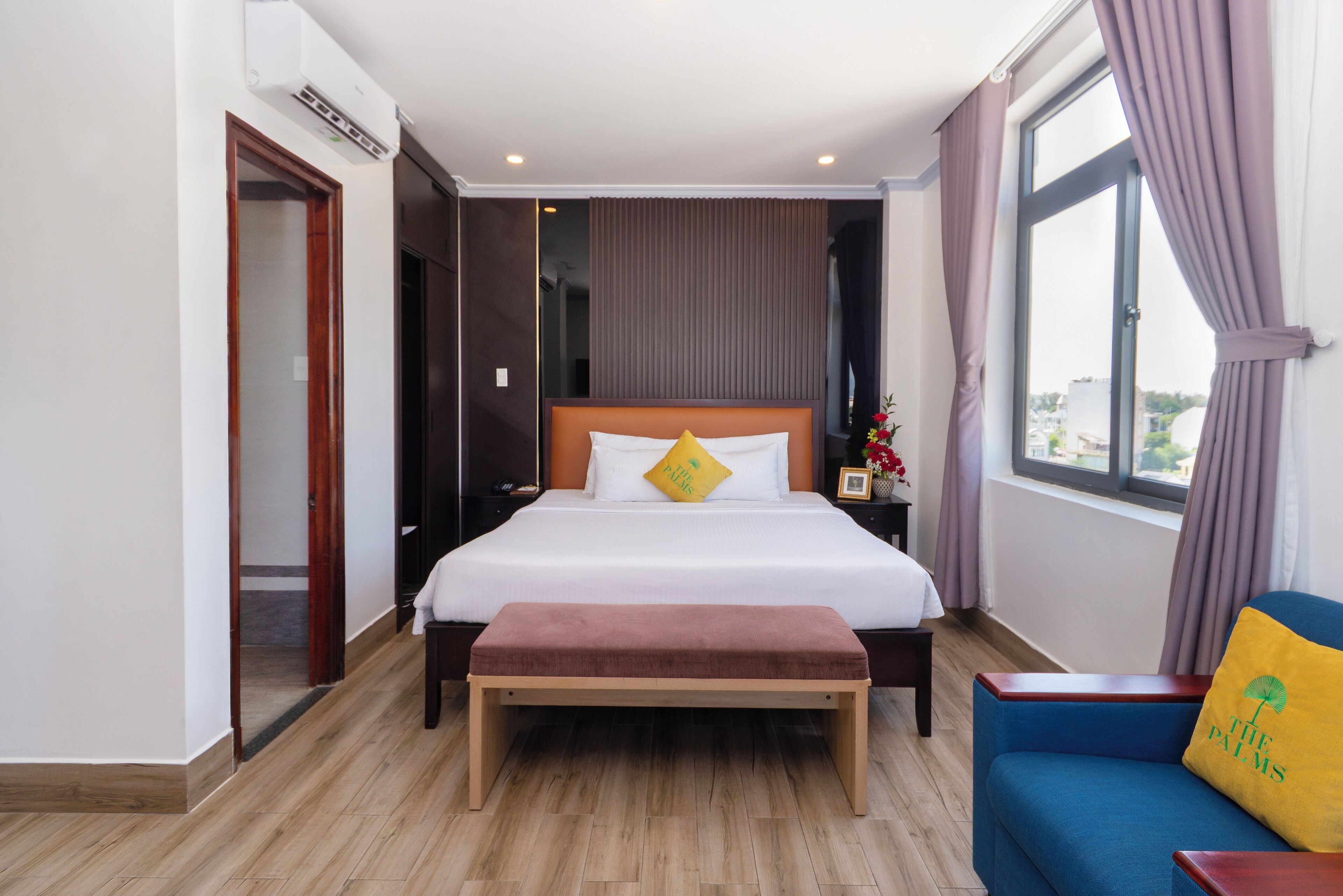 A hotel room with a large bed, a bench, a blue sofa, and a window with curtains.