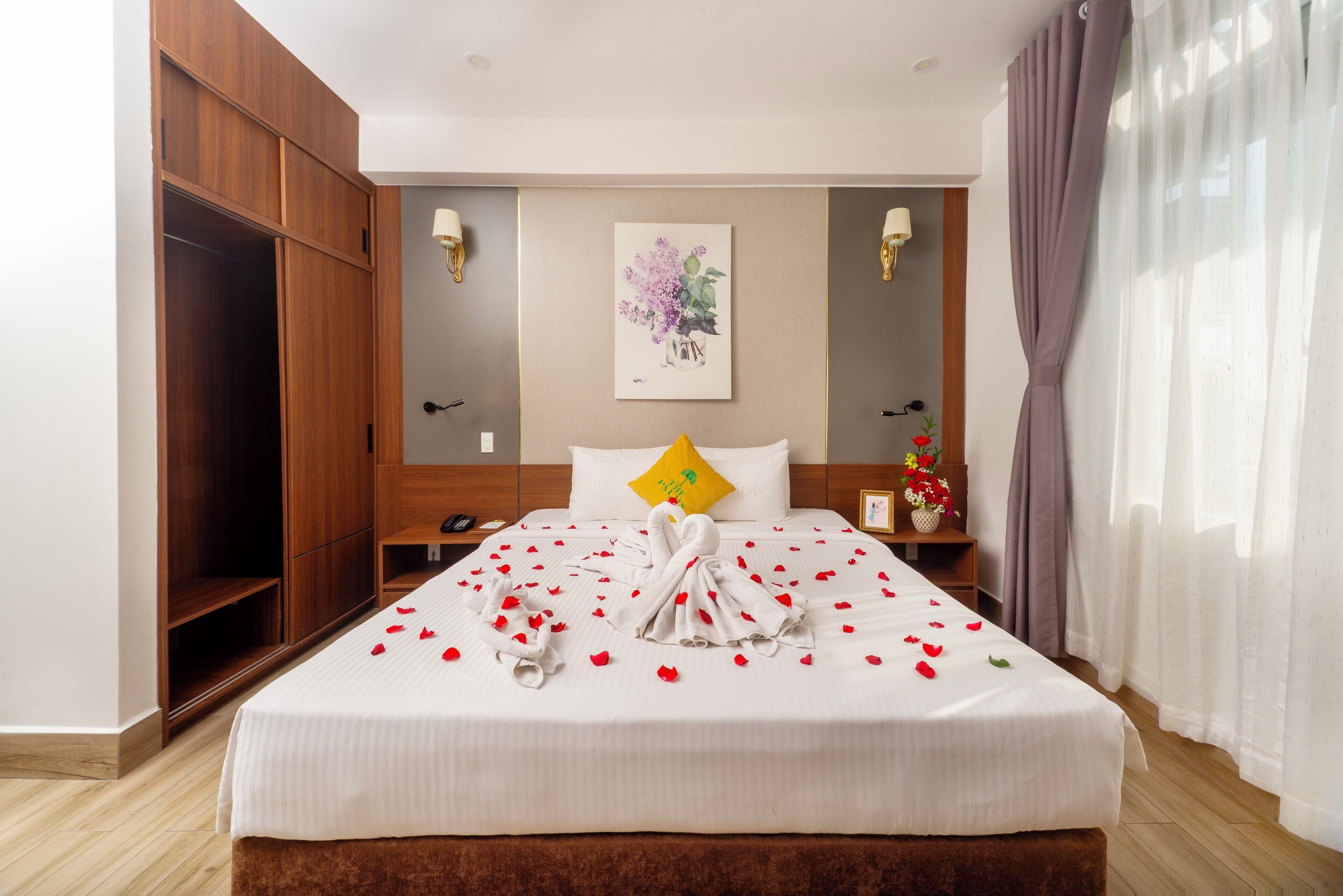 A neatly made bed with rose petals and a towel arrangement, a wooden wardrobe, and a painting on the wall.