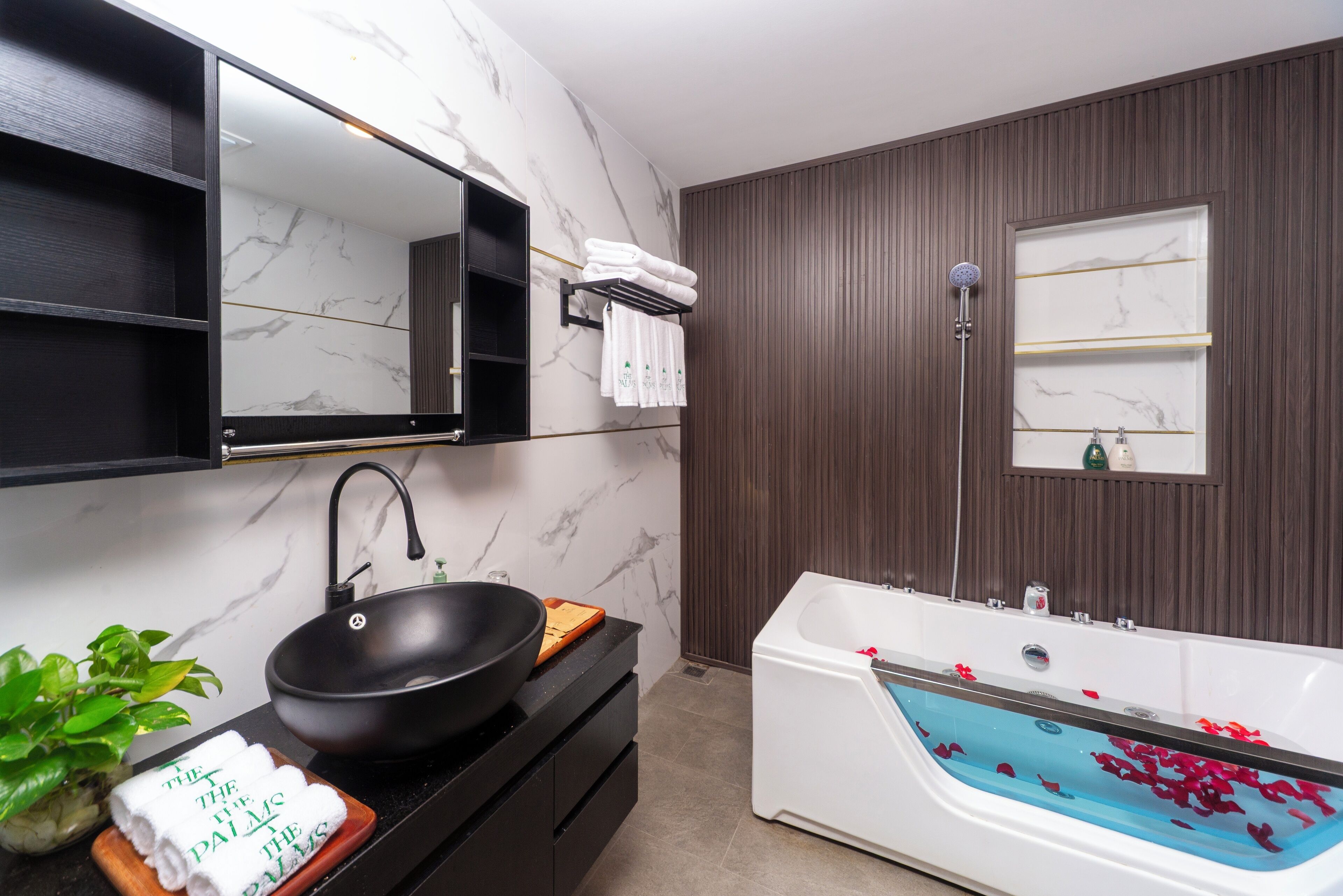 A modern bathroom with a black sink, white bathtub with rose petals, a mirror, and a potted plant.