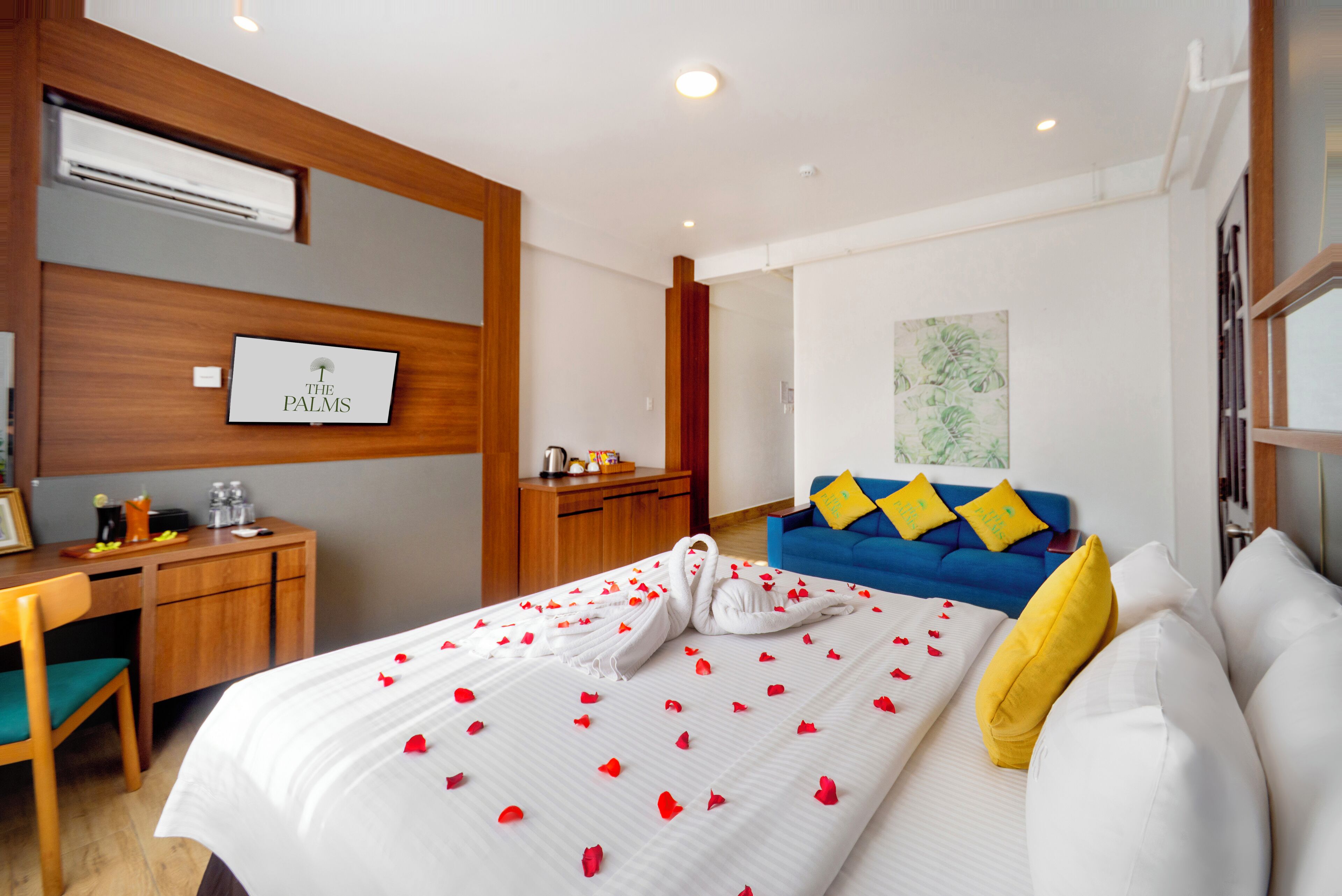 A hotel room with a bed, a desk, a blue sofa, and a wall-mounted TV displaying "THE PALMS".