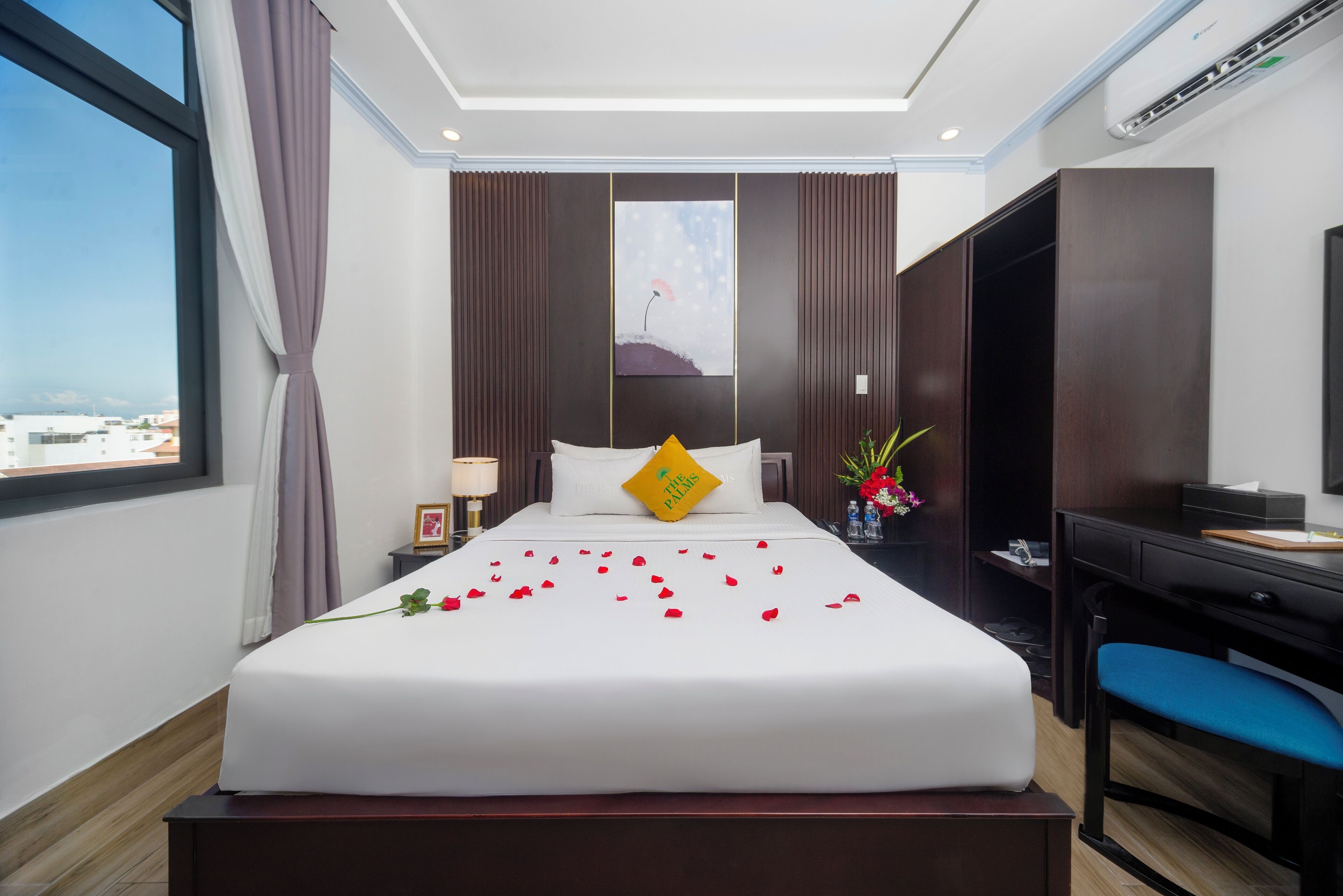 Habitación de hotel con cama, escritorio, silla, mesita de noche, lámpara, un jarrón con flores y un cuadro en la pared.