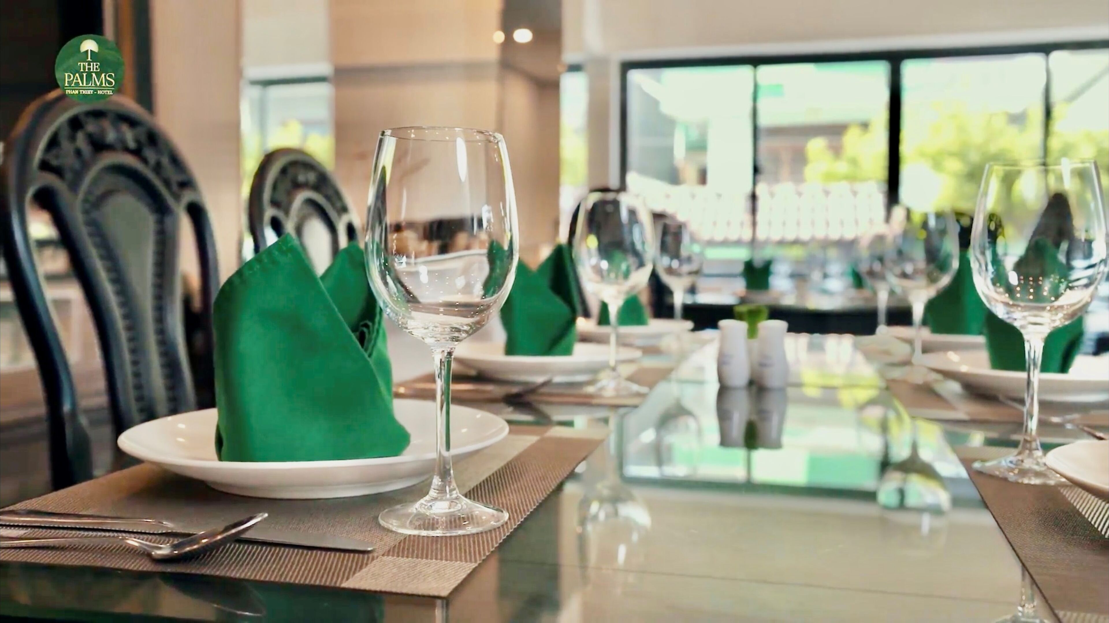 Una mesa de comedor dispuesta con copas de vino, platos y servilletas verdes, con vista a un restaurante y mesas al aire libre al fondo.