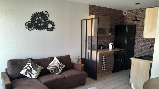 Ein kompakter Wohnbereich mit braunem Sofa, einem gemusterten Teppich, einer Küchenzeile mit Backsteinwand und einem Essbereich mit weißem Stuhl.
