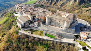 An aerial view of a historic town with a prominent castle-like structure, surrounded by terraced fields and a winding road.