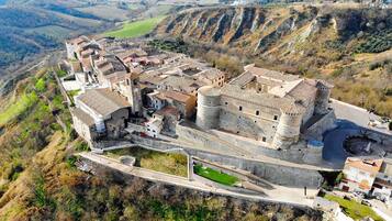 An aerial view of a historic town with a prominent castle-like structure, surrounded by terraced fields and a winding road.