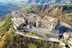 Une vue aérienne d’une ville historique, avec un imposant édifice qui ressemble à un château, entouré de champs en terrasses et d’une route sinueuse.
