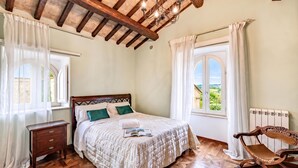 A bedroom with a wooden bed, a nightstand, a rocking chair, and a view of greenery through a window.
