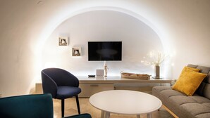 A modern living room with a flat-screen TV mounted on a white wall, a round white table, a grey sofa with a mustard-colored pillow, and a dark blue armchair. - Luan Old Town apartment by DuHomes (Dubrovnik)