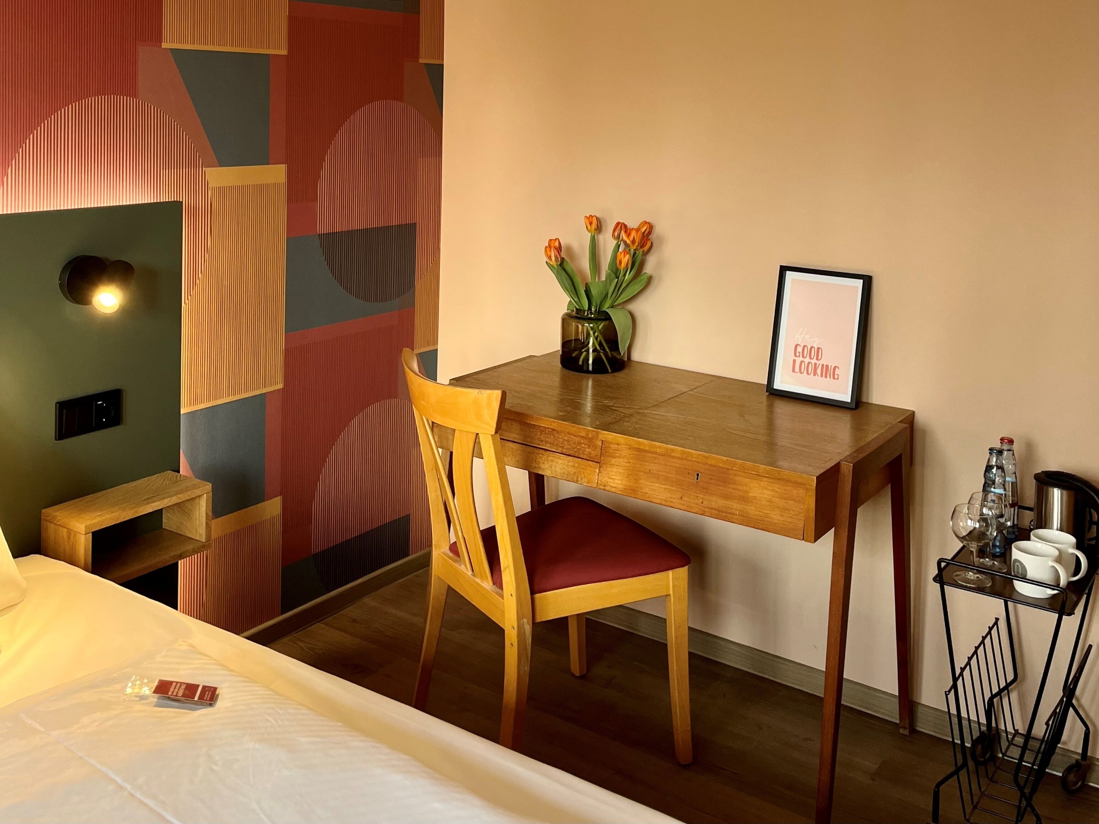 Une chambre d’hôtel de taille modeste, comprenant un bureau en bois, une chaise, une petite table de chevet, un lit, une étagère fixée au mur avec un vase de fleurs et un tableau encadré.