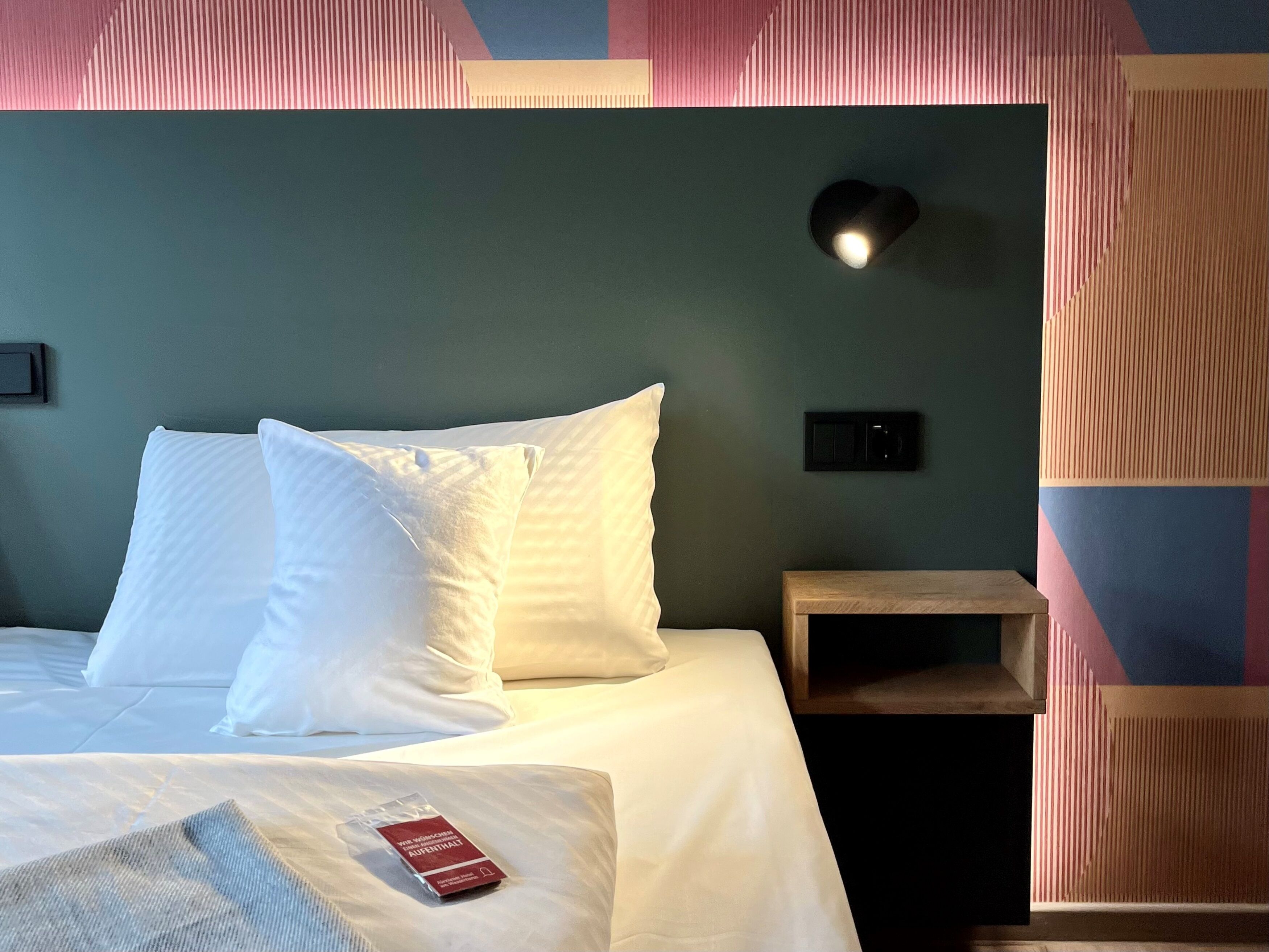 Ein ordentlich bezogenes Bett mit weißen Leinen und Kissen, ein Nachttisch mit Lampe und eine Wand mit geometrischem Muster.