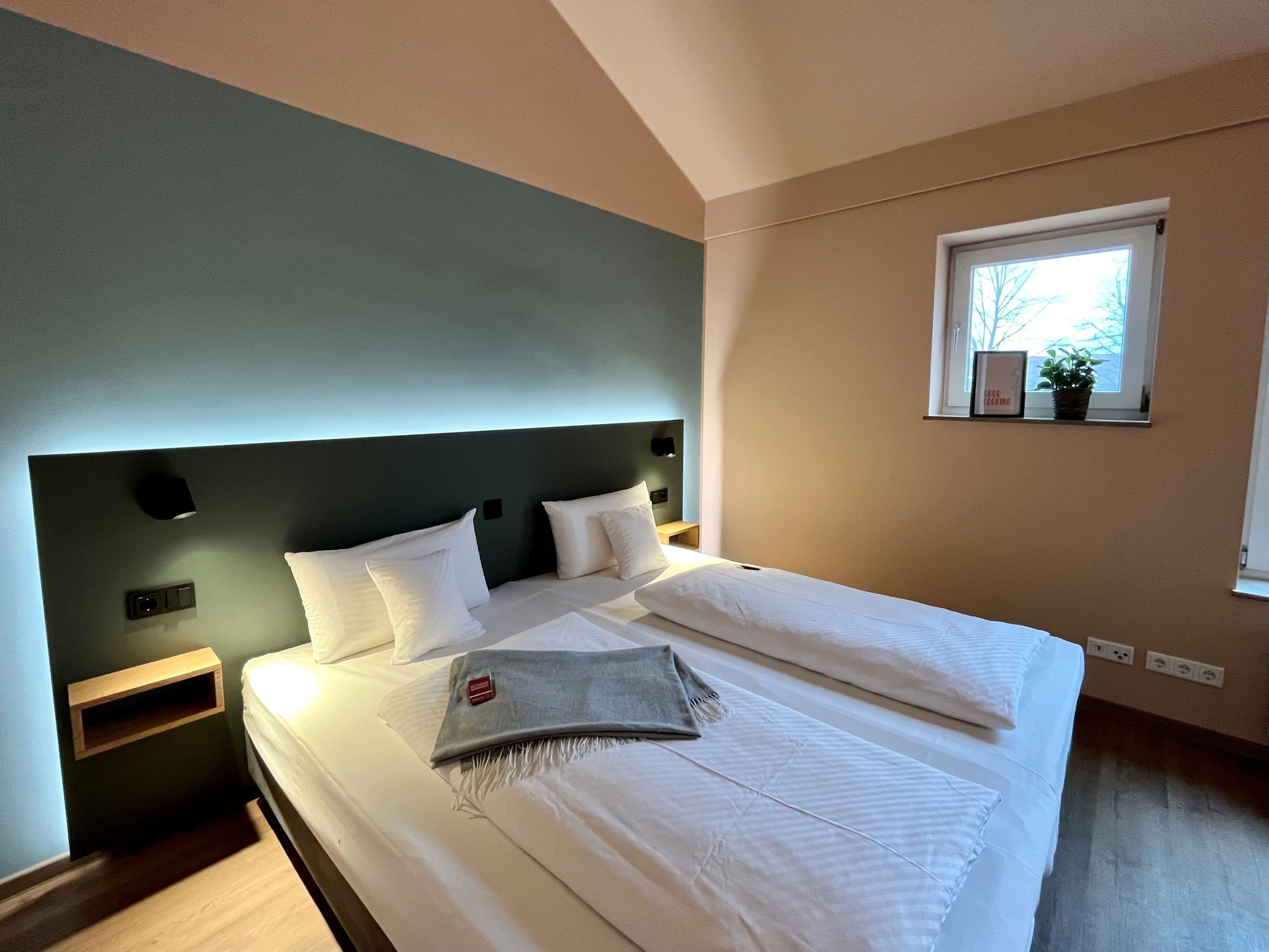 Een moderne slaapkamer met een groot bed, een hoofdbord, nachtkastjes, een raam met uitzicht en een klein plankje met een plantje.