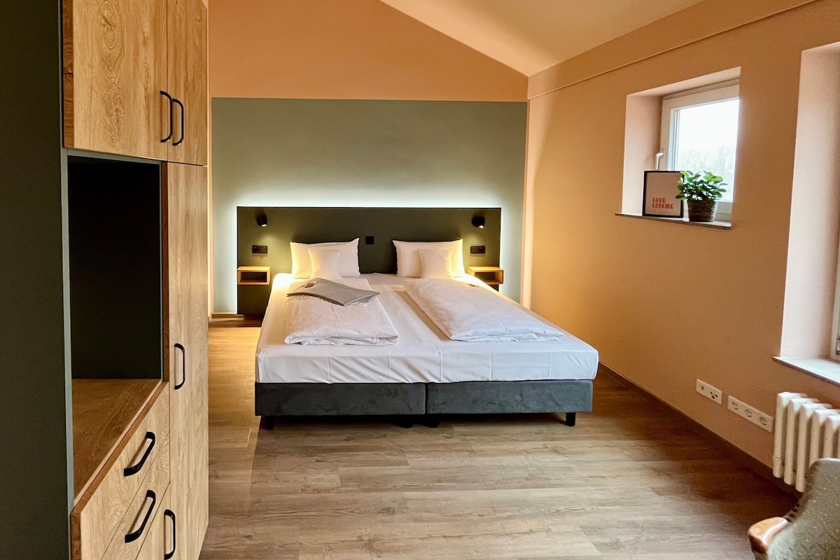 Quarto moderno com cama grande, guarda-roupa de madeira, mesas de cabeceira e janela com planta.