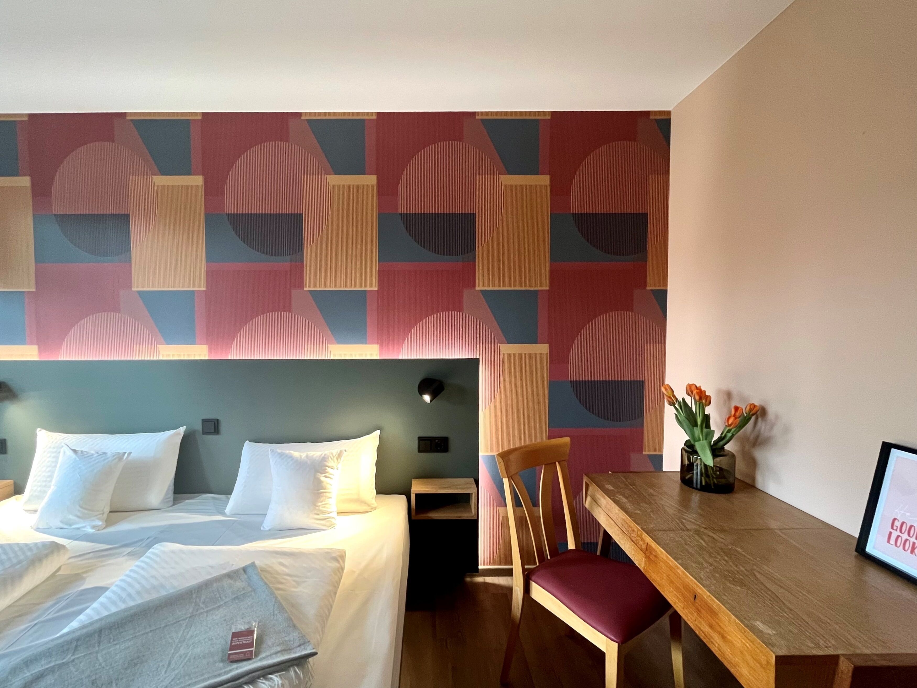 Una habitación de hotel con una cama, un escritorio de madera, una silla y una pared con patrones geométricos.