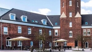 Een gebouw van baksteen met een centrale toren, meerdere ramen en buitenruimtes met parasols.