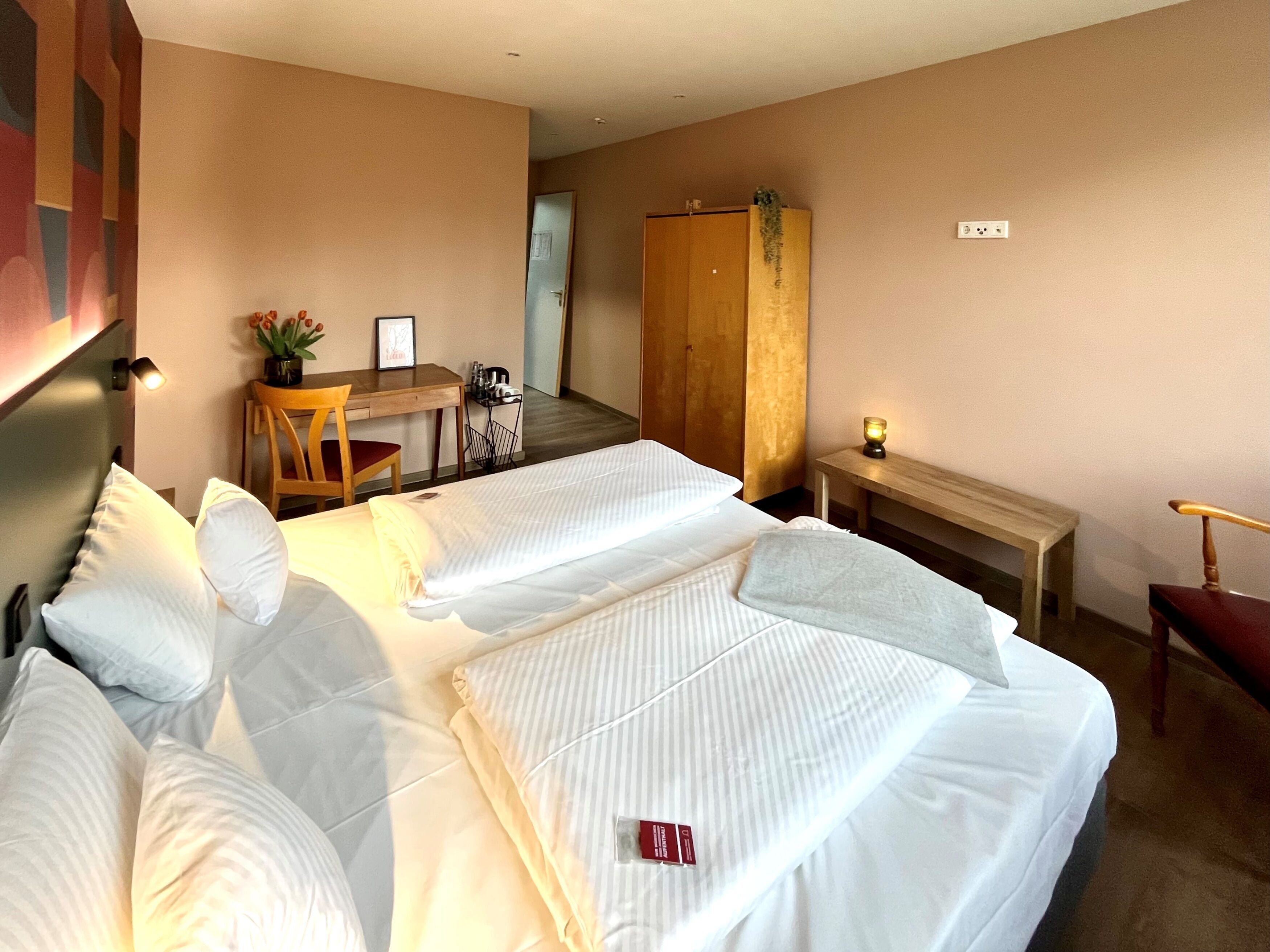 A hotel room with two beds, a desk, a chair, a wardrobe, and a small table with a lamp.