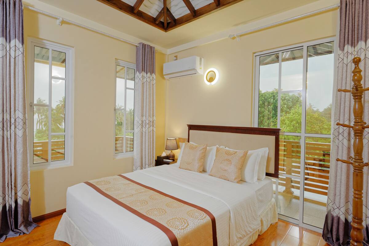 A bedroom with a large bed, a nightstand with a lamp, an air conditioning unit, and a view of the outdoors through a sliding glass door.