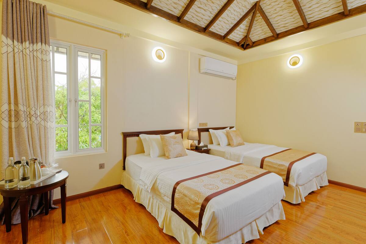 A hotel room with two beds, a wooden floor, a window with curtains, an air conditioning unit, and a table with bottles.