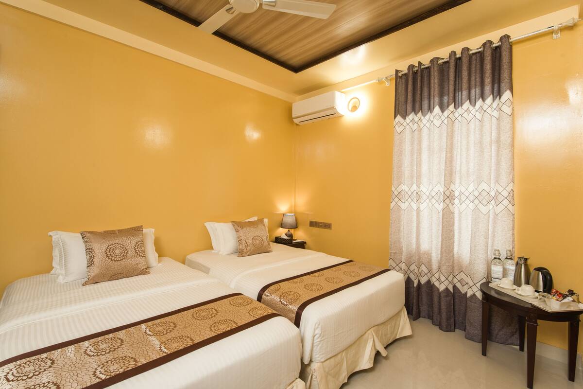 A hotel room with two beds, a ceiling fan, an air conditioning unit, a table with a kettle, and a curtain with a geometric pattern.