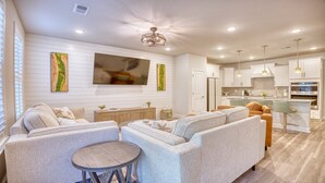 A modern living room with a large flat-screen TV, a dining area with a round table, and a kitchen with white cabinetry and a bar. - 5128 Townes at Barefoot (North Myrtle Beach)