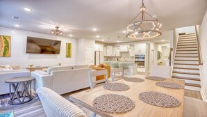 A modern living room with a sofa set, a dining table with placemats, a kitchen area, and a staircase. - 5128 Townes at Barefoot (North Myrtle Beach)