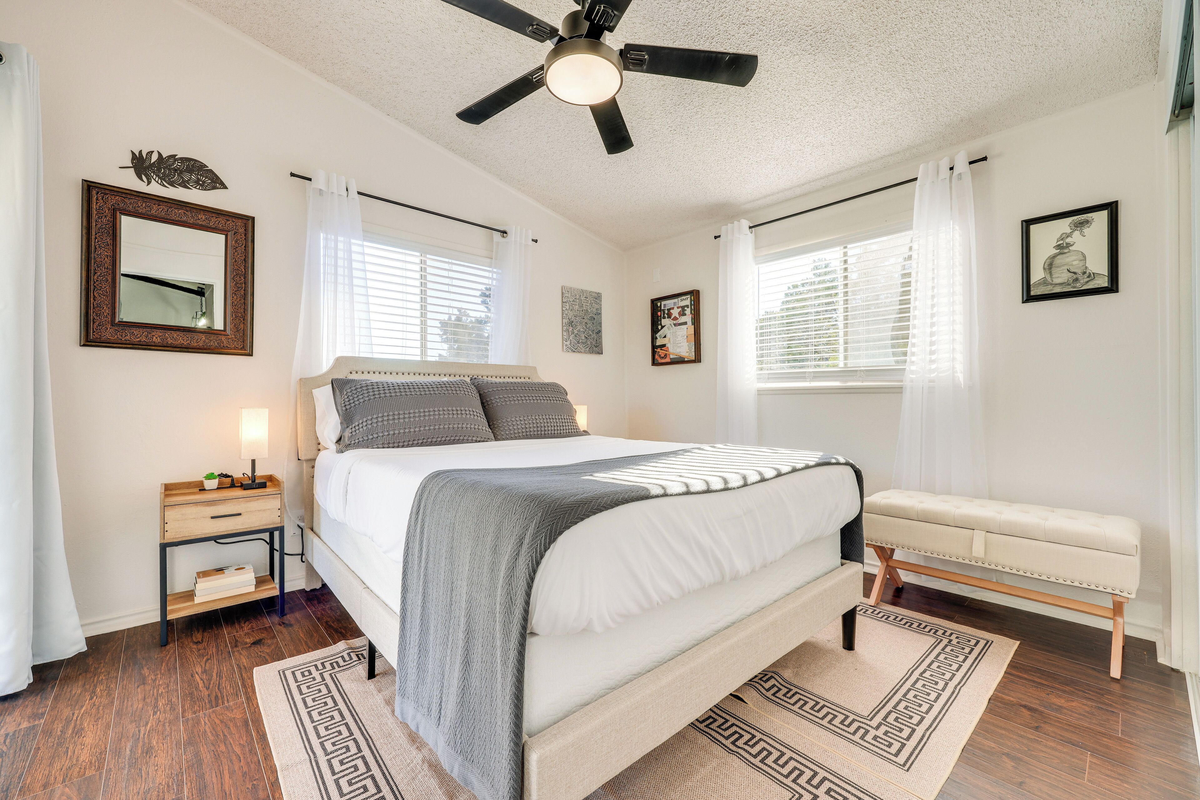 A bedroom with a bed, bedside table, bench, ceiling fan, and window with curtains.
