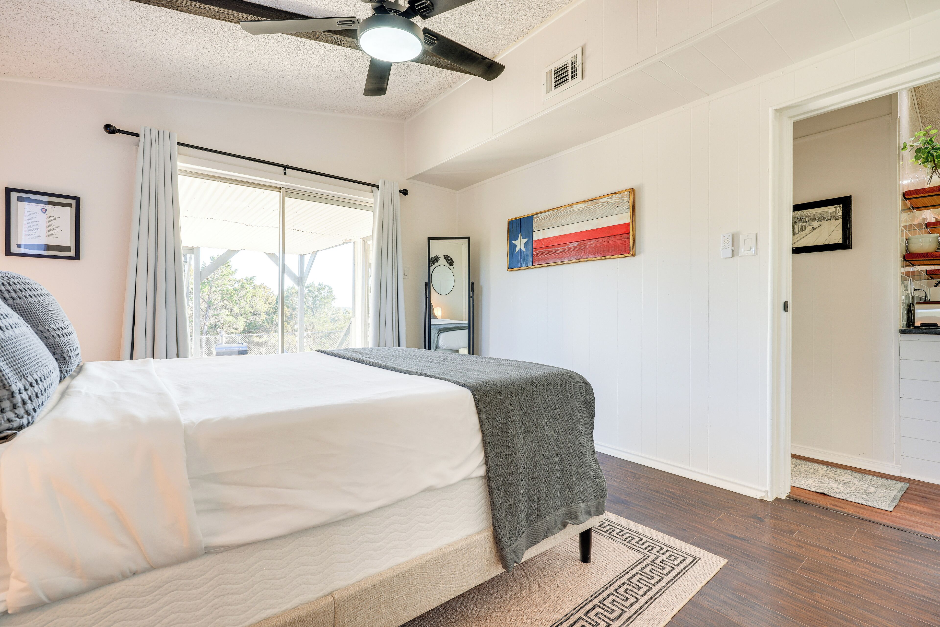 A bedroom with a bed, a ceiling fan, a window with curtains, a Texas flag, and a door leading to another room.