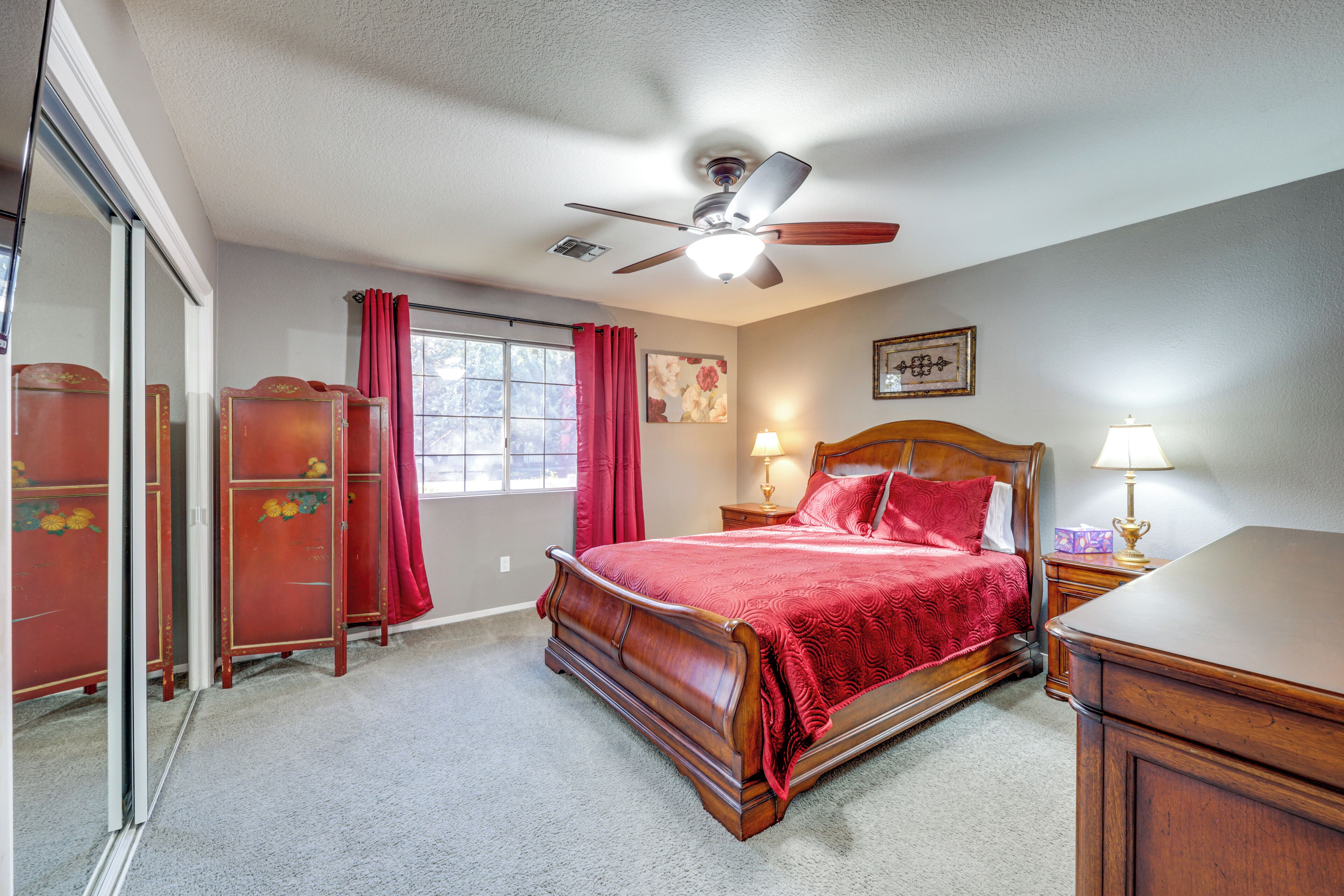 A bedroom with a wooden bed, a ceiling fan, a window with red curtains, and a nightstand with a lamp.