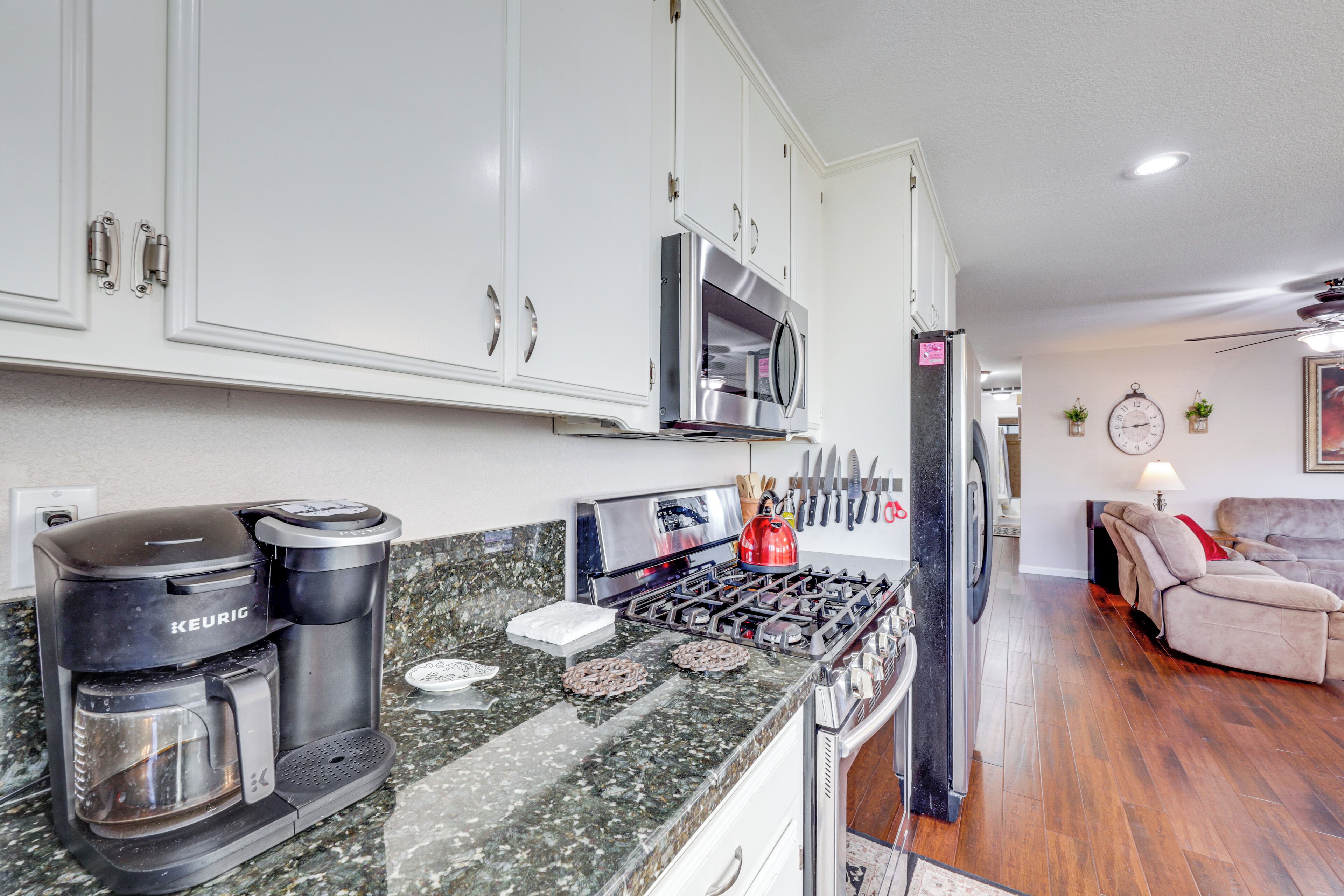 A kitchen with a Keurig coffee maker, microwave, and stove. There is a dining area with a sofa and a clock on the wall.