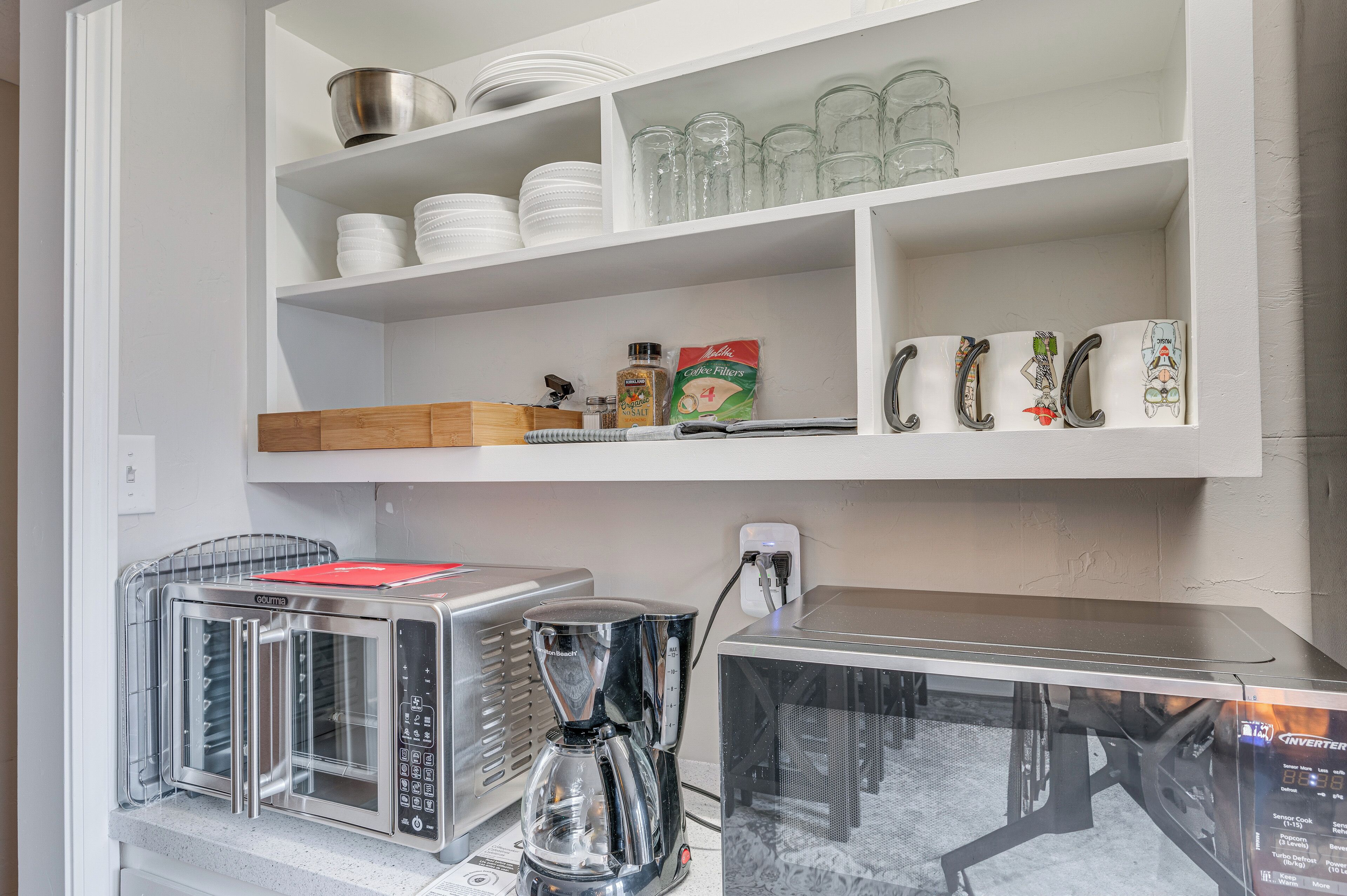 A kitchen with white shelves holding dishes, glasses, and a coffee maker. There are also two coffee machines and a refrigerator.