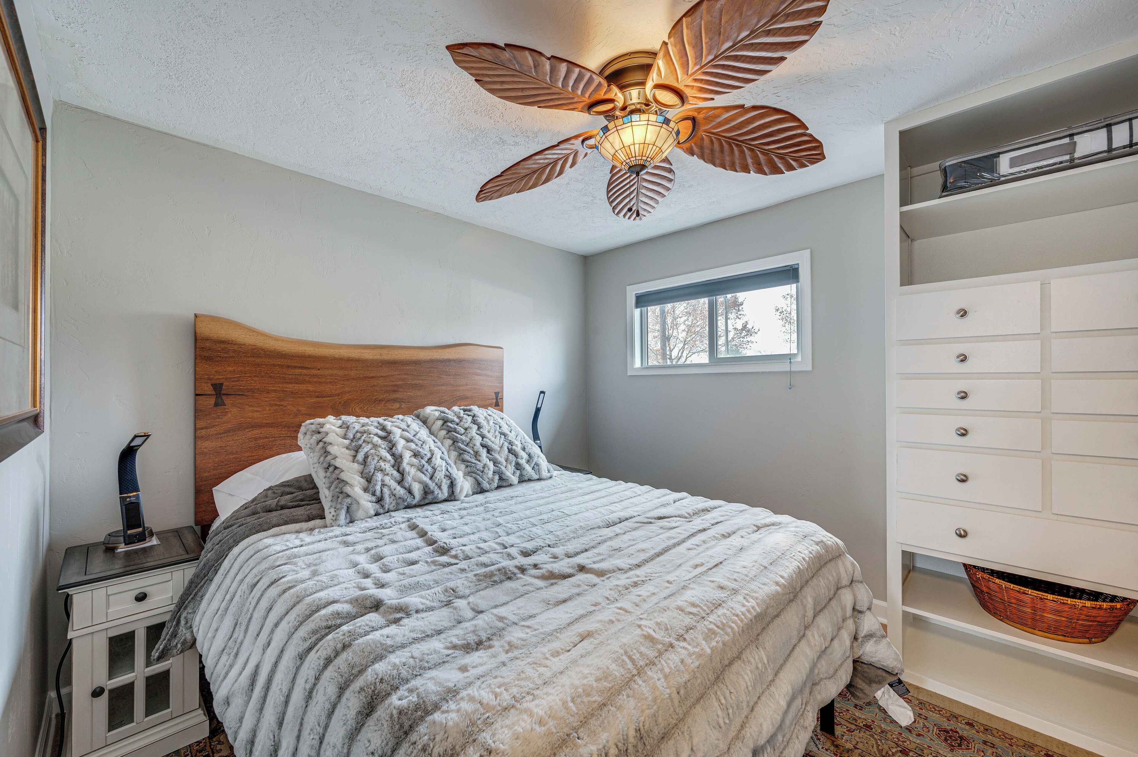 A bedroom with a wooden headboard, a bed with a white and gray blanket, a white dresser, and a ceiling fan.
