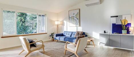 Een moderne woonkamer met een blauwe bank, twee fauteuils, een houten vloer, een groot raam en een wandairconditioner.