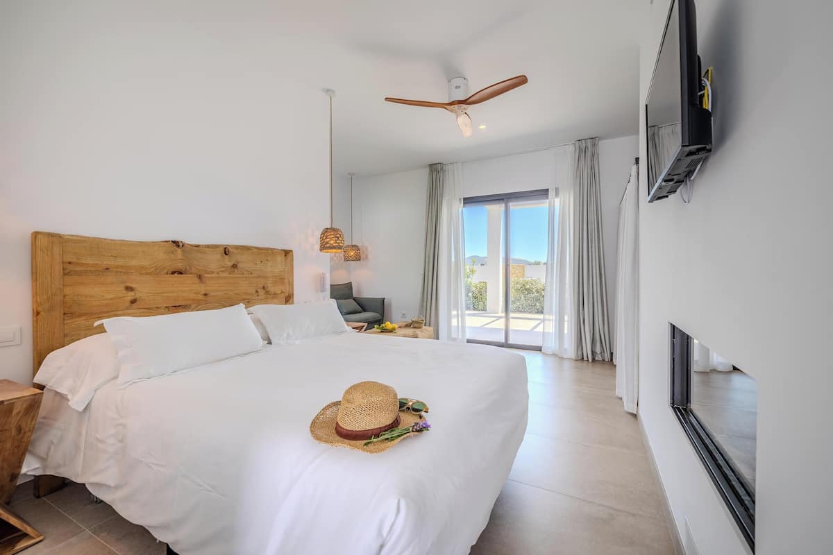 Quarto de hotel com cabeceira de madeira, cama com lençóis brancos, chapéu de palha, óculos de sol e um buquê sobre a cama. Há um ventilador de teto, uma TV fixada na parede e vista para o exterior através de uma porta de correr de vidro.
