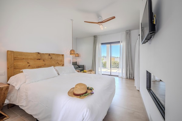 Habitación de hotel con cabecera de madera, cama con ropa blanca, un sombrero de paja, gafas de sol y un ramo sobre la cama. Hay un ventilador de techo, un televisor montado en la pared y vista al exterior a través de una puerta corrediza de vidrio.