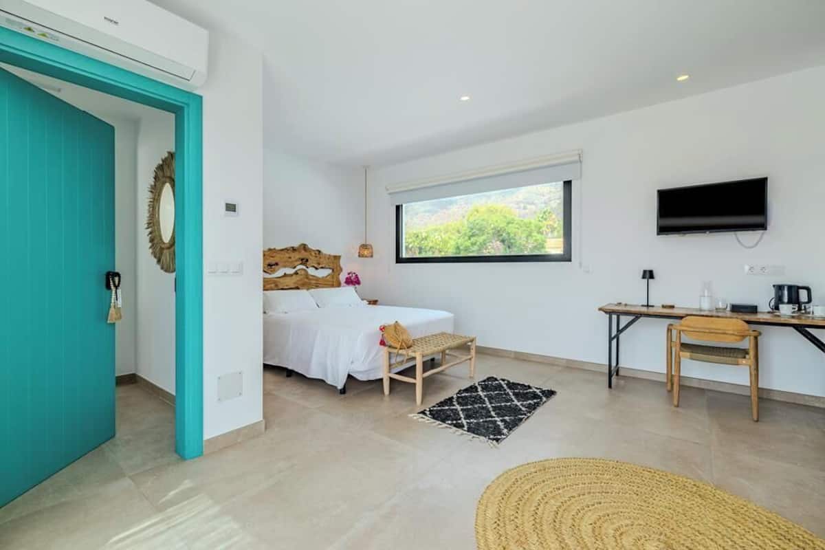 Quarto com cama, escrivaninha, televisão e janela com vista para árvores.