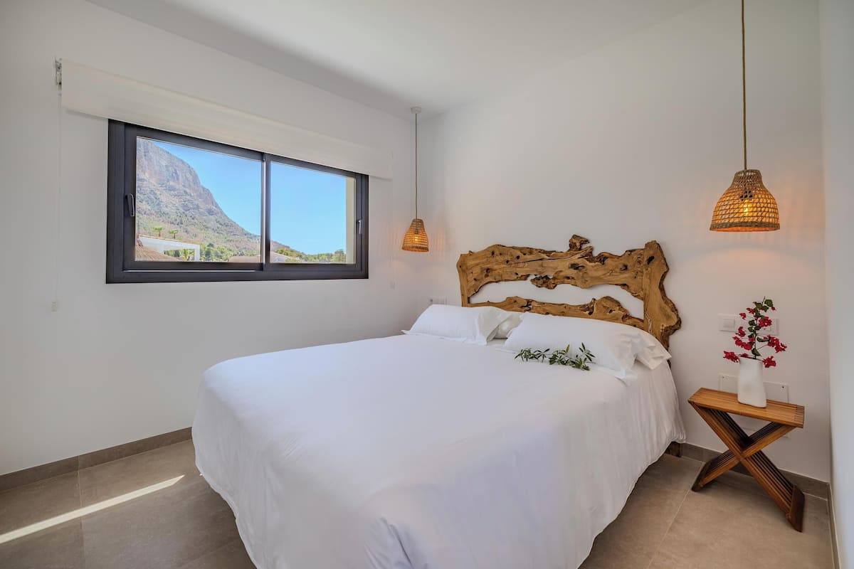 Quarto com cama grande, cabeceira de madeira, janela com vista para as montanhas e duas luminárias pendentes.