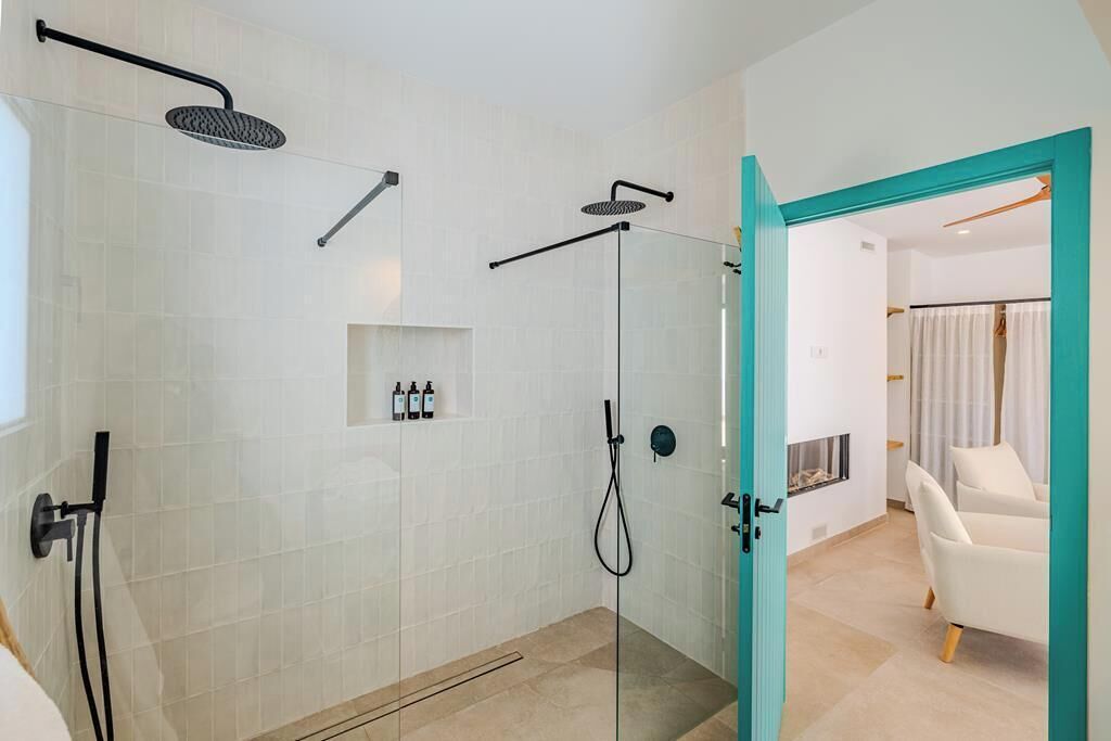 A modern bathroom with a glass shower enclosure, a white tiled wall, and a teal-framed door leading to a living area with a white sofa.