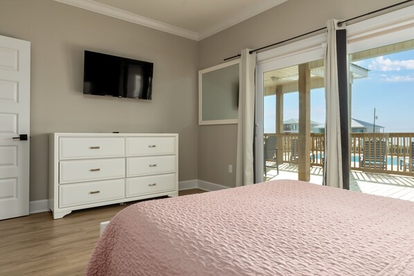 Un dormitorio con cama, cómoda blanca, televisor y puerta corrediza de vidrio a un balcón con piscina.