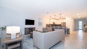 A modern living room with a sofa, a TV, and a kitchen in the background. - 9056sh-the Retreat At Championsgate 8 Bedroom Home by RedAwning (Davenport)