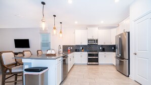 A modern kitchen with white cabinets, stainless steel appliances, and a central island with seating. - 9056sh-the Retreat At Championsgate 8 Bedroom Home by RedAwning (Davenport)