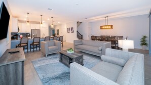 A modern living room with a grey sofa, a coffee table, and a TV on a wooden stand. There is a kitchen area with white cabinets and a dining area with a table and chairs. - 9056sh-the Retreat At Championsgate 8 Bedroom Home by RedAwning (Davenport)