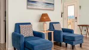 Una habitación con dos sillones azules, una mesita con una lámpara, un cuadro del océano y un cartel que dice "BEACH HOUSE".