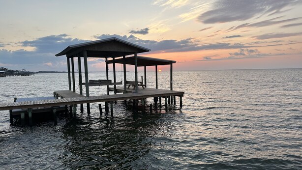 Eine Holzstege, der sich über ruhiges Wasser erstreckt, mit überdachter Sitzgelegenheit und Blick auf den Sonnenuntergang.