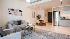 A modern living room with a sofa, coffee table, and dining area. A kitchen with a refrigerator and microwave is visible in the background. - Aya - Beachfront Apartment With all Amenities and Pool (Dubai)